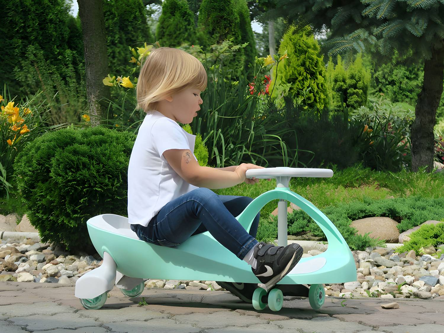 Un niño monta un coche de juguete verde azulado y blanco en un camino pavimentado. El fondo presenta arbustos verdes, árboles y flores amarillas.