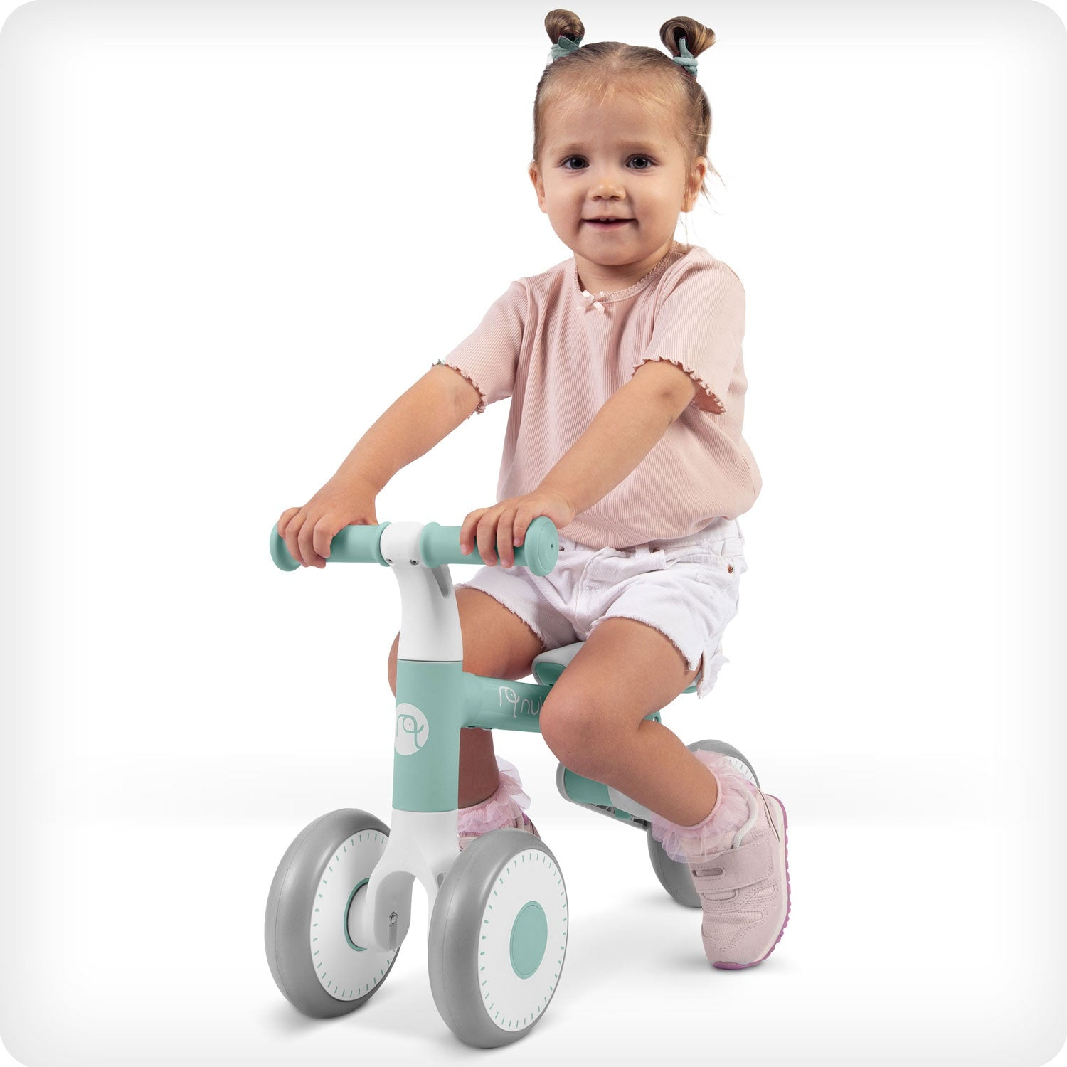 Una niña con coletas sonríe mientras está sentada en una bicicleta de equilibrio verde y blanca. Lleva top rosa, pantalones cortos blancos y zapatos rosas.