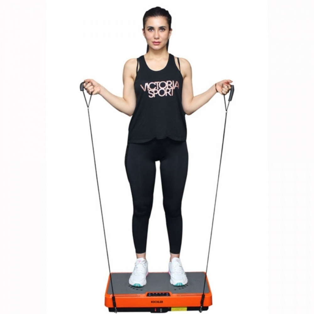 Spor giyimli bir kadın, titreşimli bir egzersiz platformunda duruyor, tutma kolları tutuyor. Siyah üst ve tayt, beyaz ayakkabılar giyiyor.