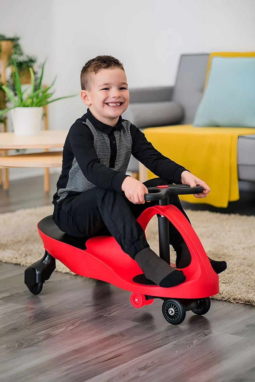 Un niño sonriente monta un coche de juguete rojo. Lleva ropa negra. El fondo muestra una sala de estar con un sofá y plantas.