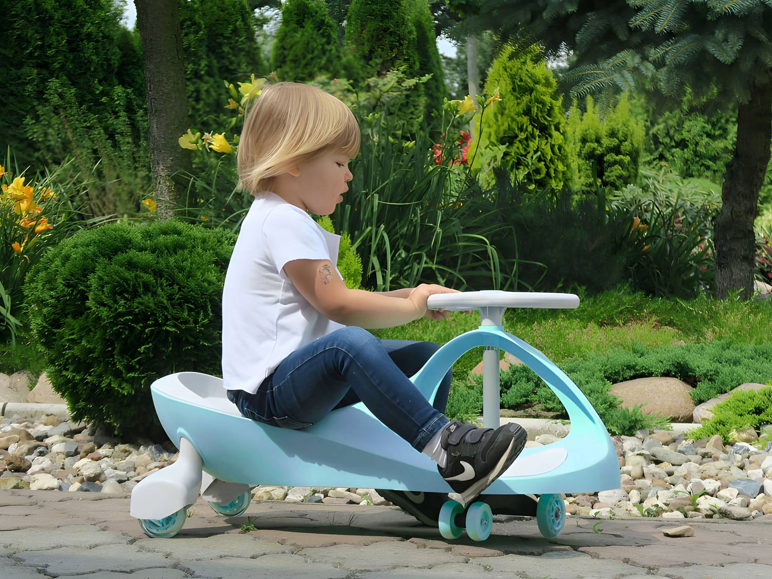 Un niño monta un coche de juguete azul al aire libre. El niño lleva una camisa blanca y vaqueros. Árboles y arbustos verdes llenan el fondo.