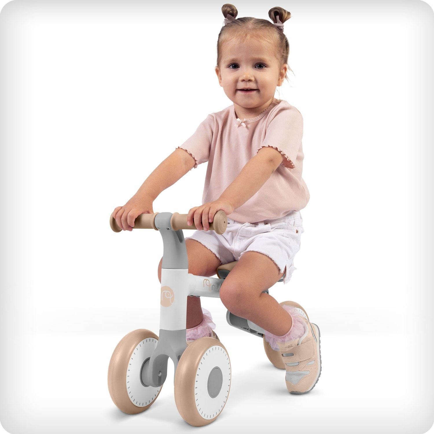 Una niña pequeña monta en una bicicleta de equilibrio. Lleva una camisa rosa y pantalones cortos blancos. La bicicleta es gris y beige. Fondo blanco.