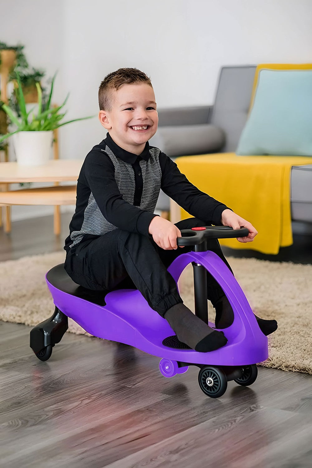 Un niño sonriente monta un coche de juguete morado en el interior. Viste de negro y está frente a un sofá con una manta amarilla.