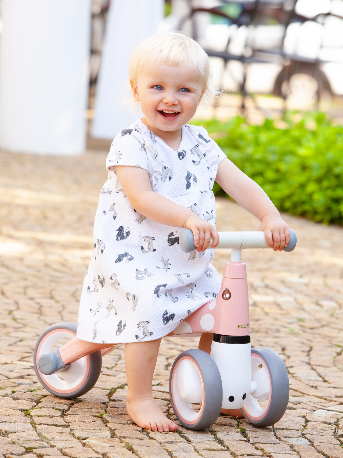 Un bambino sorridente con un vestito bianco su una bici senza pedali rosa. Il bambino ha i capelli biondi ed è a piedi nudi. Lo sfondo è sfocato.