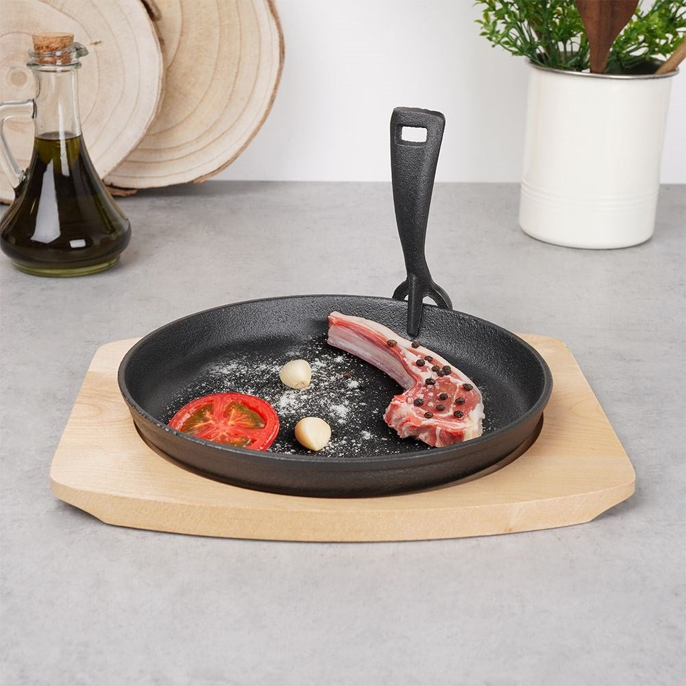 Eine Bratpfanne mit Fleisch, Tomate, Knoblauch auf einem Holzbrett. Ölflasche und Pflanze im Hintergrund.