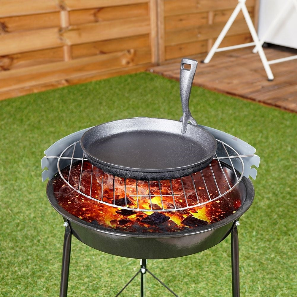 Eine schwarze gusseiserne Pfanne steht auf einem Grill über heißen Kohlen in einem Holzkohlegrill. Grünes Gras und Holzzaun sind im Hintergrund.
