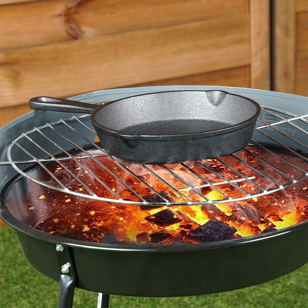 Eine kleine schwarze Gusseisenpfanne steht auf einem Grill über heißer Kohle in einem schwarzen Grill. Holzzaun im Hintergrund.
