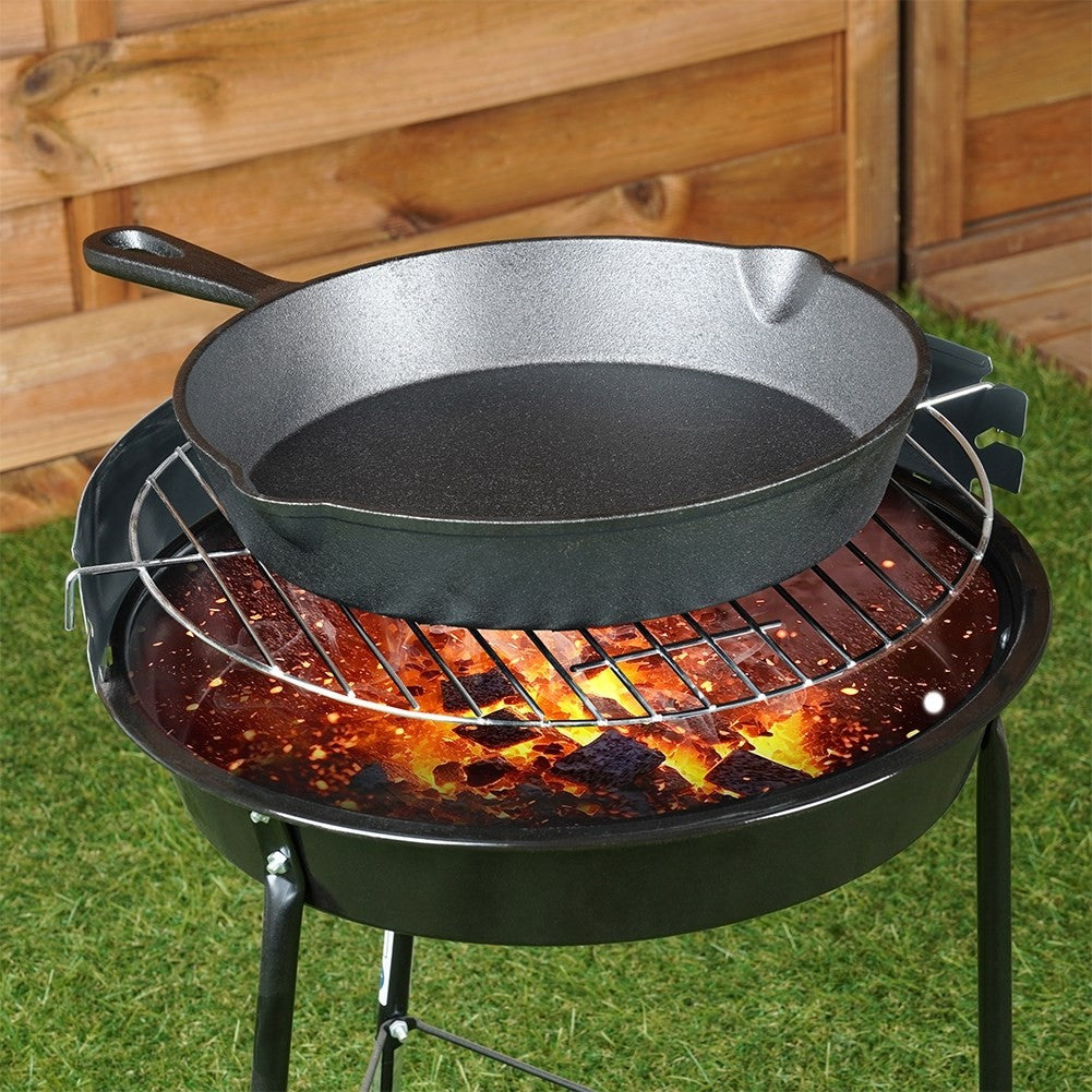 Eine schwarze Gusseisenpfanne steht auf einem Grill über einem Grill mit glühenden Glut. Der Hintergrund zeigt Holzpaneele und Gras.