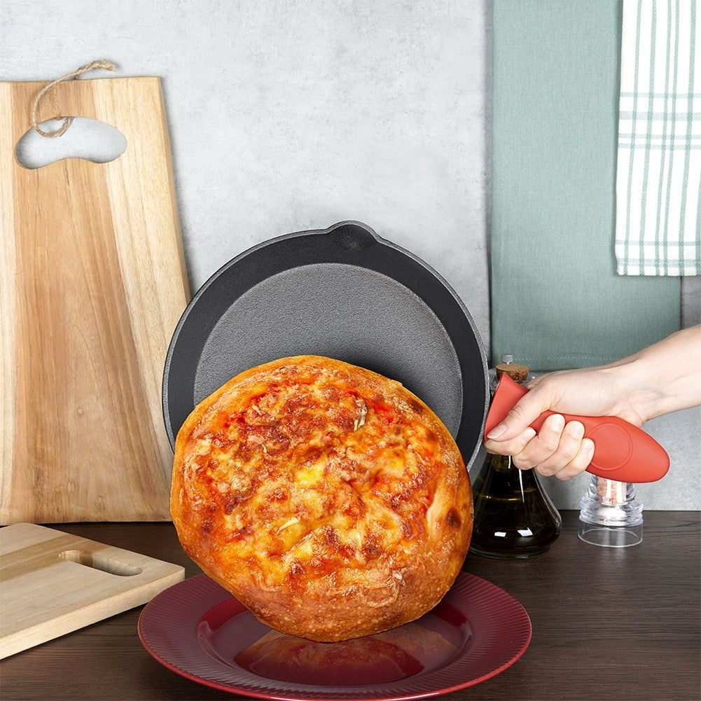 Eine gebackene Pizza in einer gusseisernen Pfanne auf einem roten Teller. Eine Hand hält die Pfanne mit rotem Griff. Holzschneidebretter im Hintergrund.