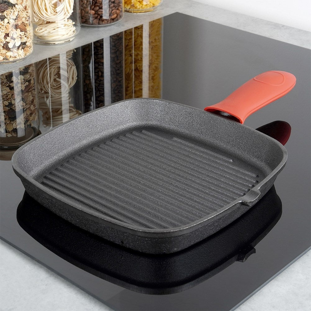Eine schwarze Grillpfanne mit orangefarbenem Griff steht auf einem schwarzen Herd. Gläser mit Lebensmitteln im Hintergrund.