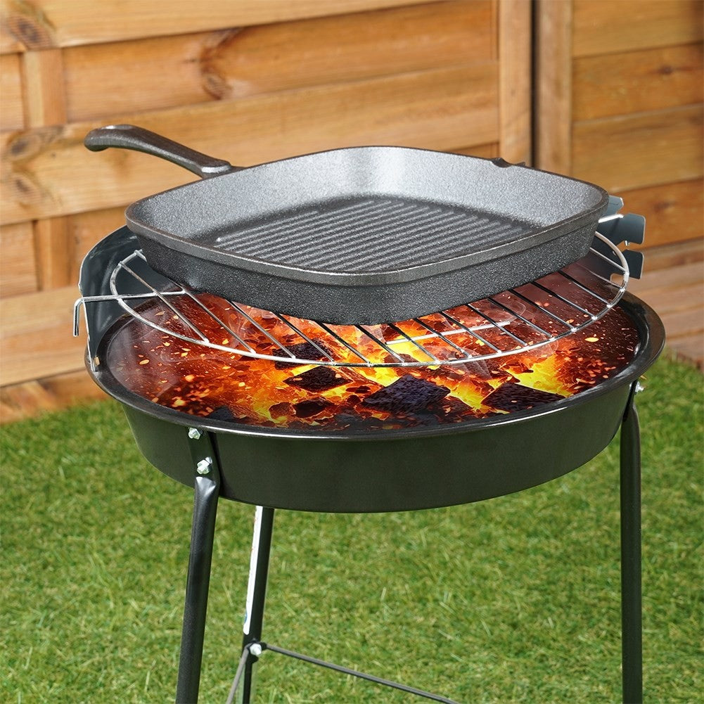 Ein Holzkohlegrill mit Grillpfanne. Glühende Kohlen sichtbar. Auf grünem Gras mit Holzzaun im Hintergrund.