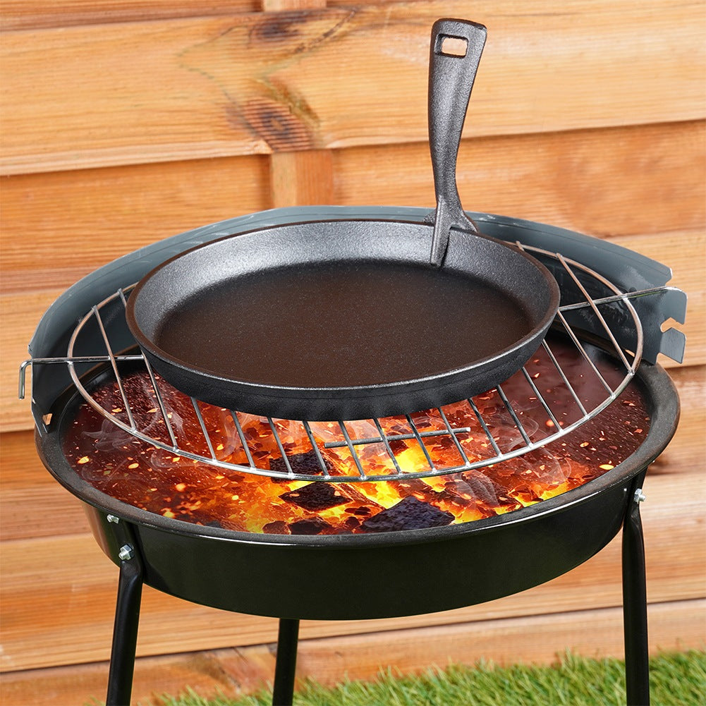 Eine schwarze Gusseisenpfanne steht auf einem Grill über heißen Kohlen in einem schwarzen Holzkohlegrill. Brauner Holzzaun im Hintergrund.