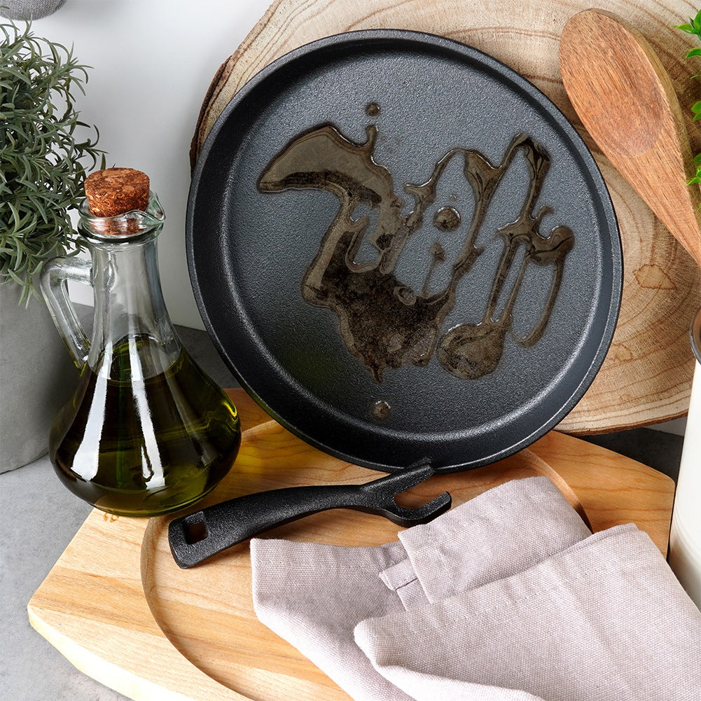 Eine schwarze Pfanne mit Öl, eine Glasflasche mit Öl, ein Holzlöffel, ein schwarzes Utensil und ein Leinentuch liegen auf einem Holzbrett.