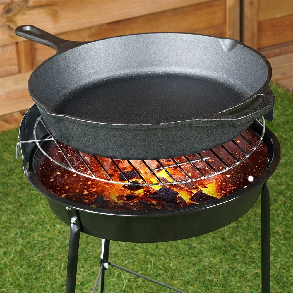 Eine schwarze Gusseisenpfanne auf einem Grill mit glühenden Kohlen. Der Grill steht auf Rasen mit Holzzaun im Hintergrund.