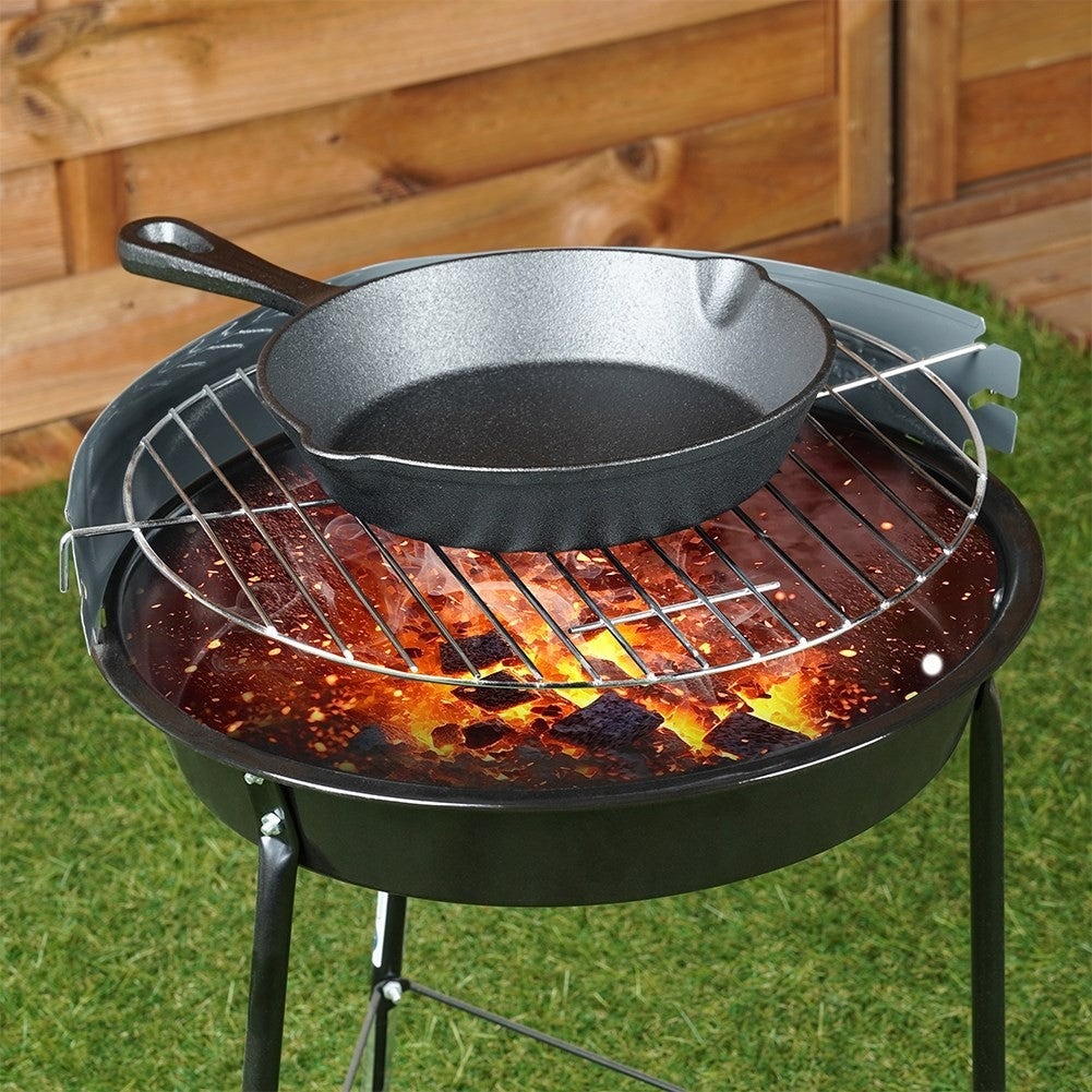 Eine schwarze Gusseisenpfanne auf einem Grill mit Feuer. Der Grill steht auf einem grünen Rasen mit einem Holzzaun im Hintergrund.
