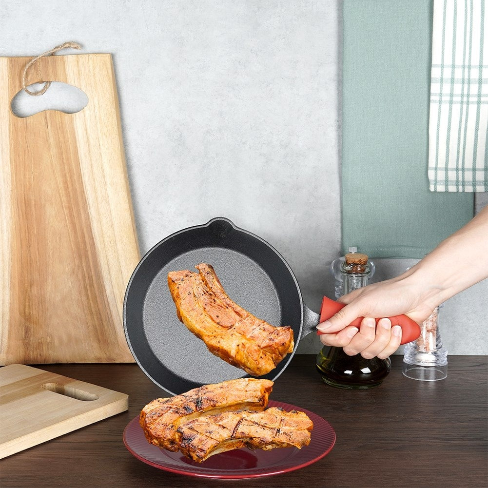 Eine Hand hält eine Pfanne mit gebratenem Fleisch. Zwei Stücke gebratenes Fleisch liegen auf einem roten Teller. Holzzuschneidebretter sind im Hintergrund.