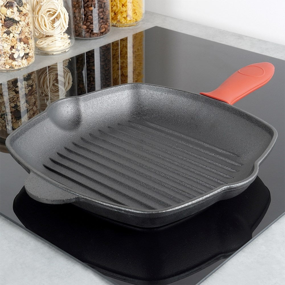 Eine schwarze Grillpfanne mit orangefarbenem Griff steht auf einem schwarzen Herd. Gläser mit Pasta und Getreide sind im Hintergrund.