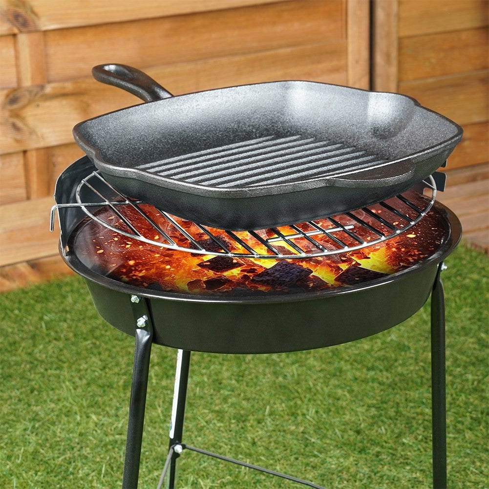 Ein schwarzer Grill mit einer gusseisernen Pfanne. Flammen sind unter dem Grill zu sehen. Gras und Holzzaun im Hintergrund.