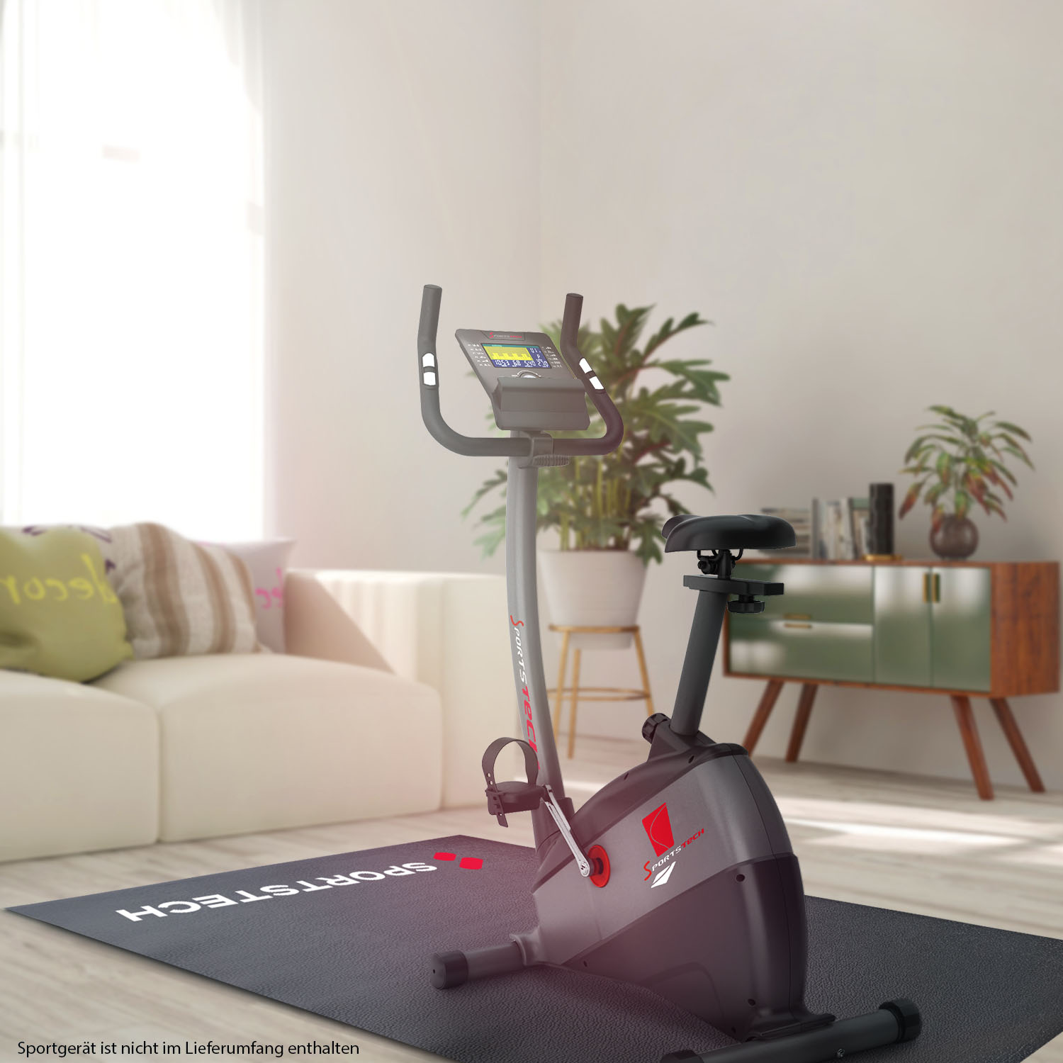 Ein Heimtrainer steht auf einer schwarzen Matte. Im Hintergrund befindet sich ein beiges Sofa. Auf der rechten Seite befinden sich ein Schrank und Pflanzen.