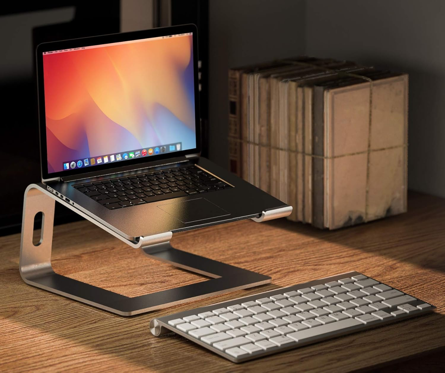 Laptop auf einem Ständer, Tastatur und Bücherstapel auf einem Holztisch. Laptop-Bildschirm zeigt farbenfrohes Wallpaper.