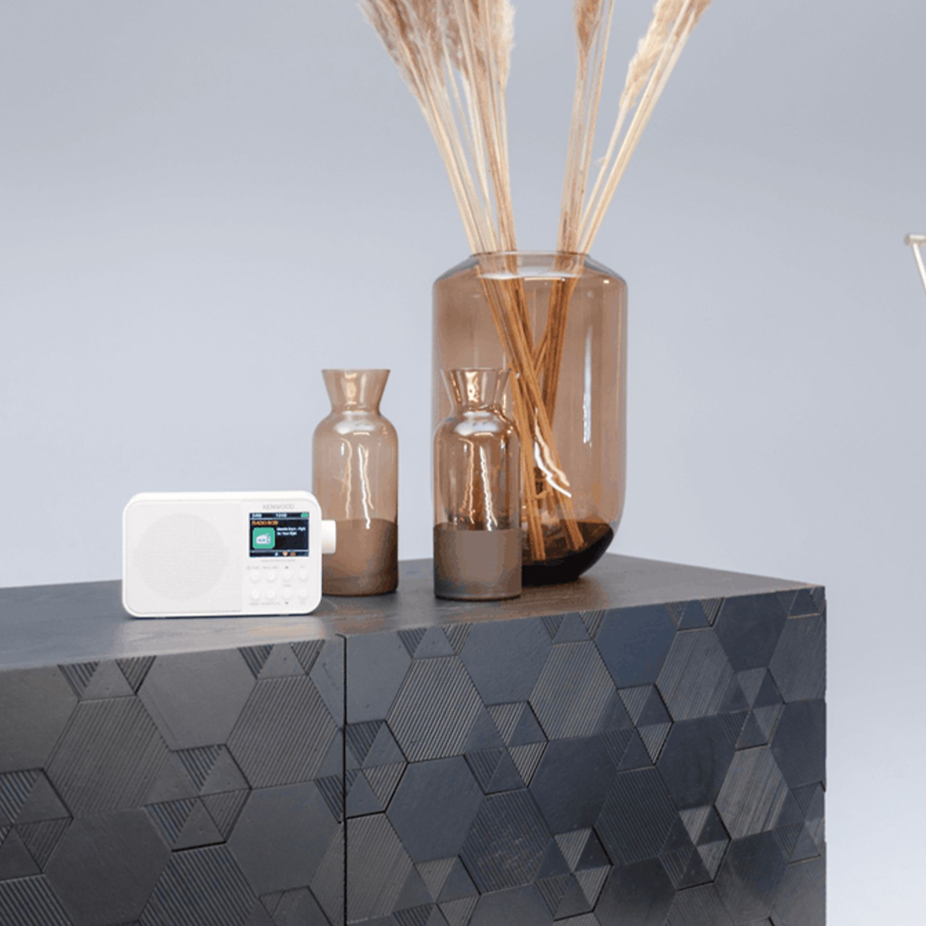 Białe radio stoi na ciemnej szafce z brązowymi wazonami i wysoką trawą. Tło jest jasnoszare.