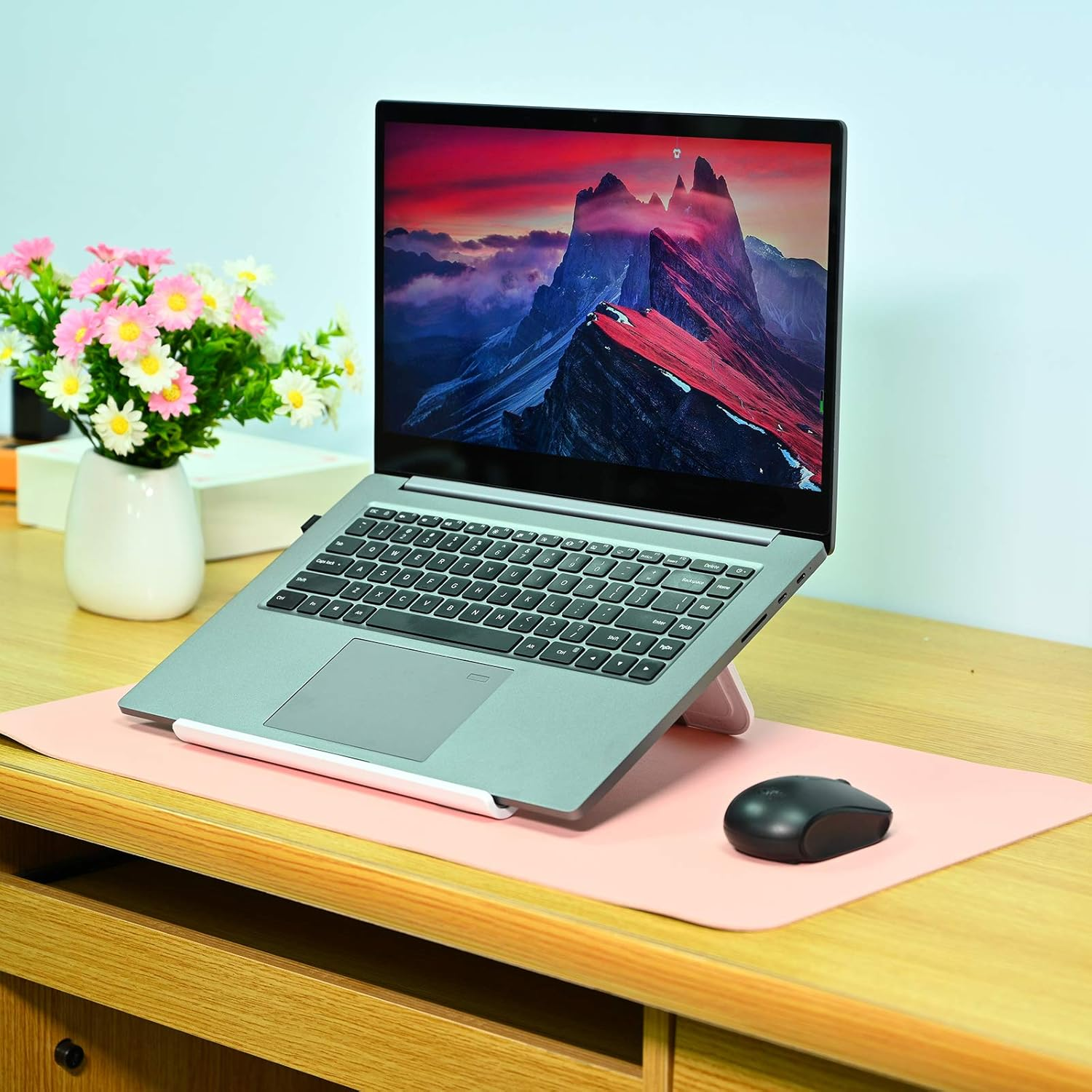 Laptop auf einem Ständer, Maus und eine rosa Matte auf einem Holztisch. Blumen in einer Vase sind links. Der Laptop zeigt eine Berglandschaft.
