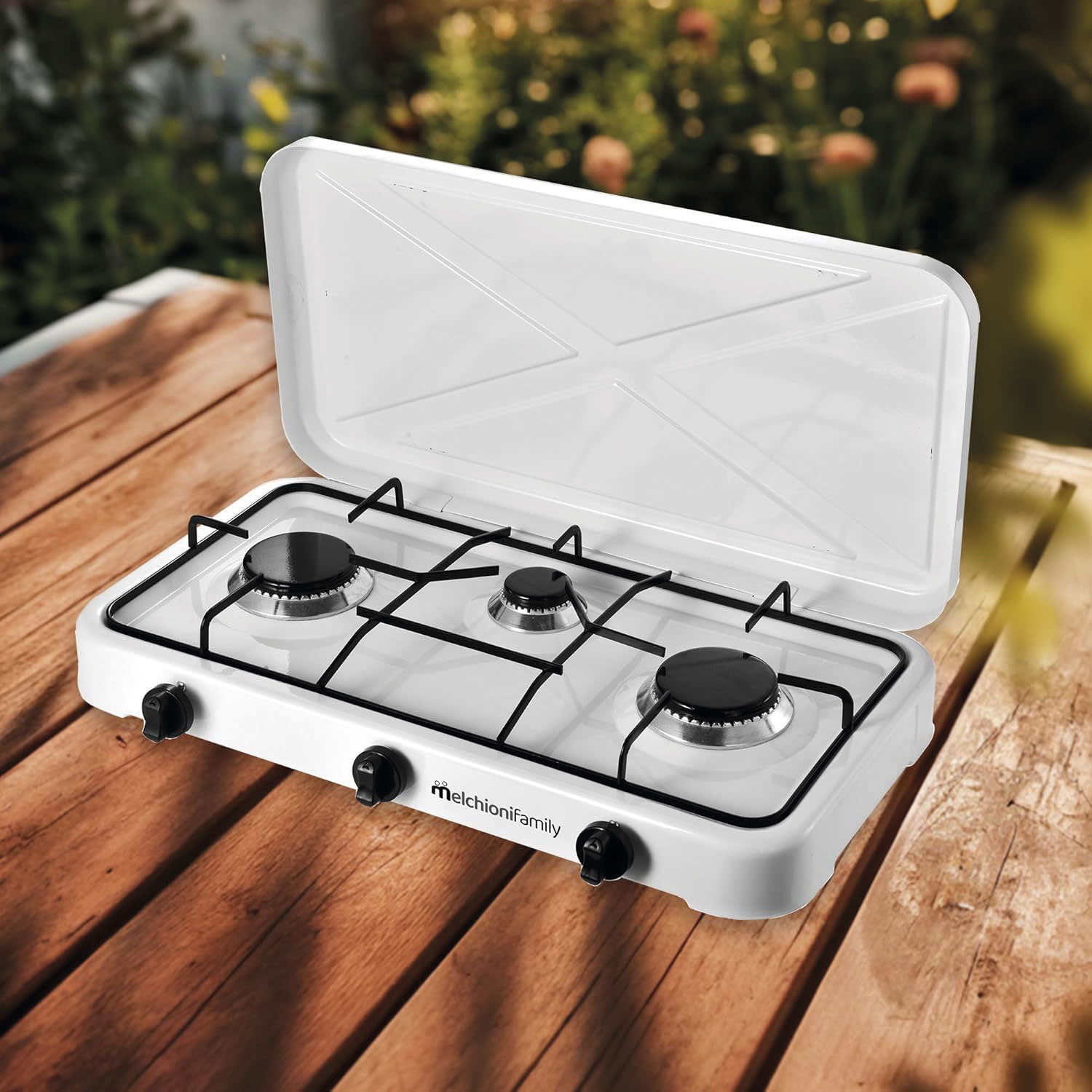 Una estufa de gas portátil blanca con tres quemadores está sobre una mesa de madera. La tapa está abierta. Hay follaje verde al fondo.