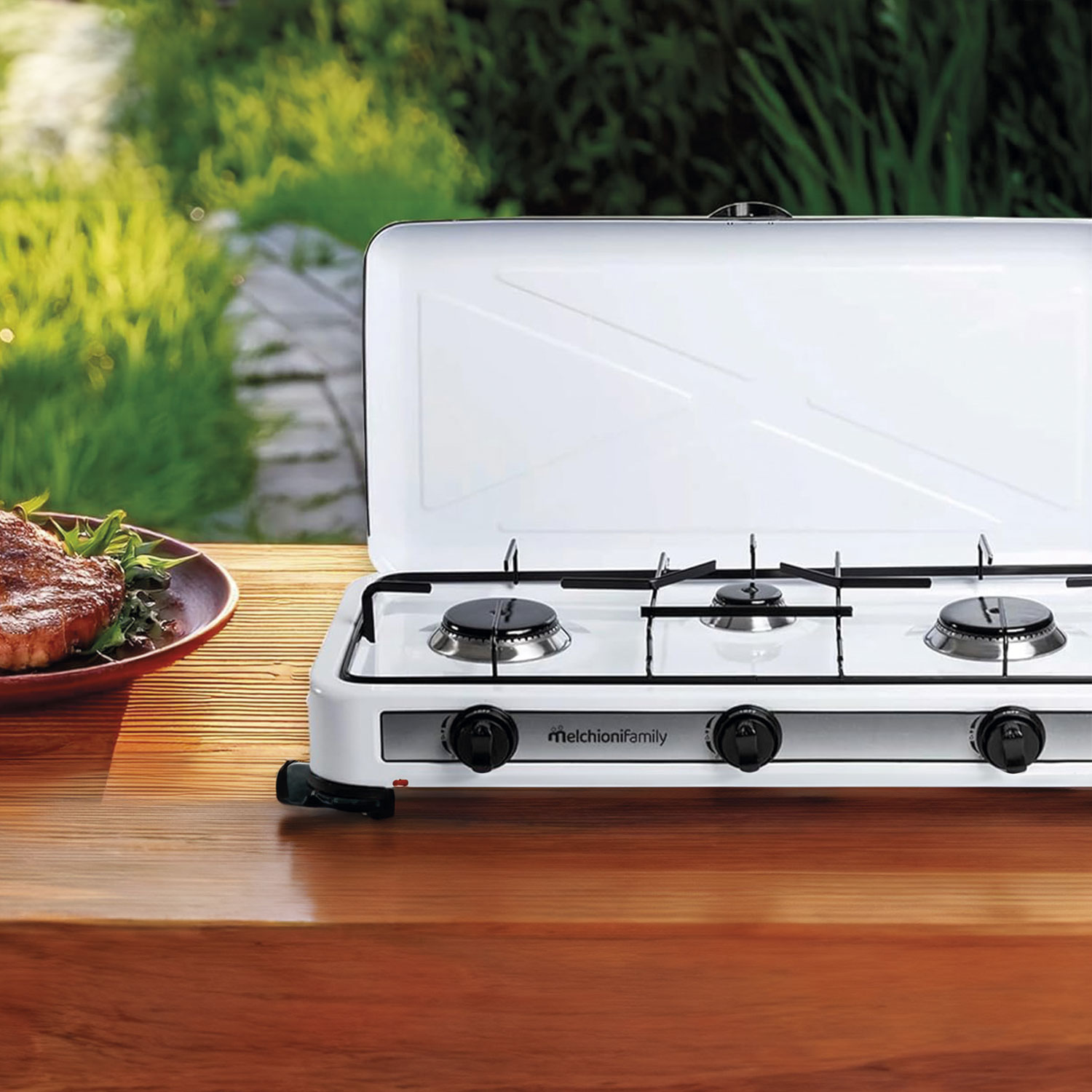 Una estufa de gas blanca con tapa está sobre una superficie de madera. Un plato con bistec y verduras está a la izquierda. Follaje verde en el fondo.