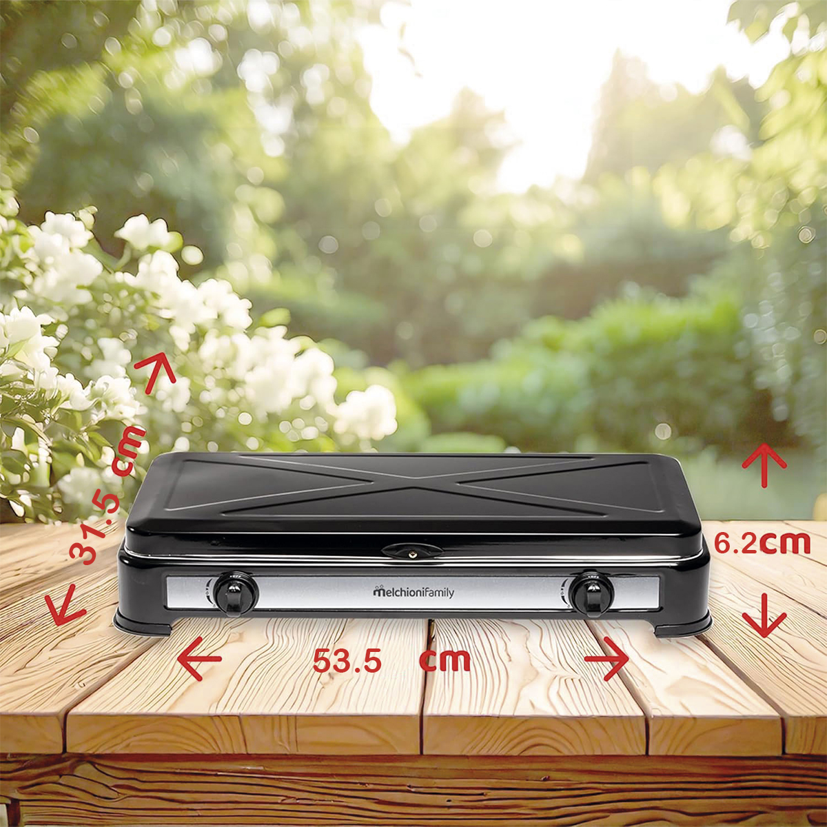 Parrilla portátil negra sobre una mesa de madera con dimensiones etiquetadas. El fondo muestra un jardín con flores blancas y follaje verde.
