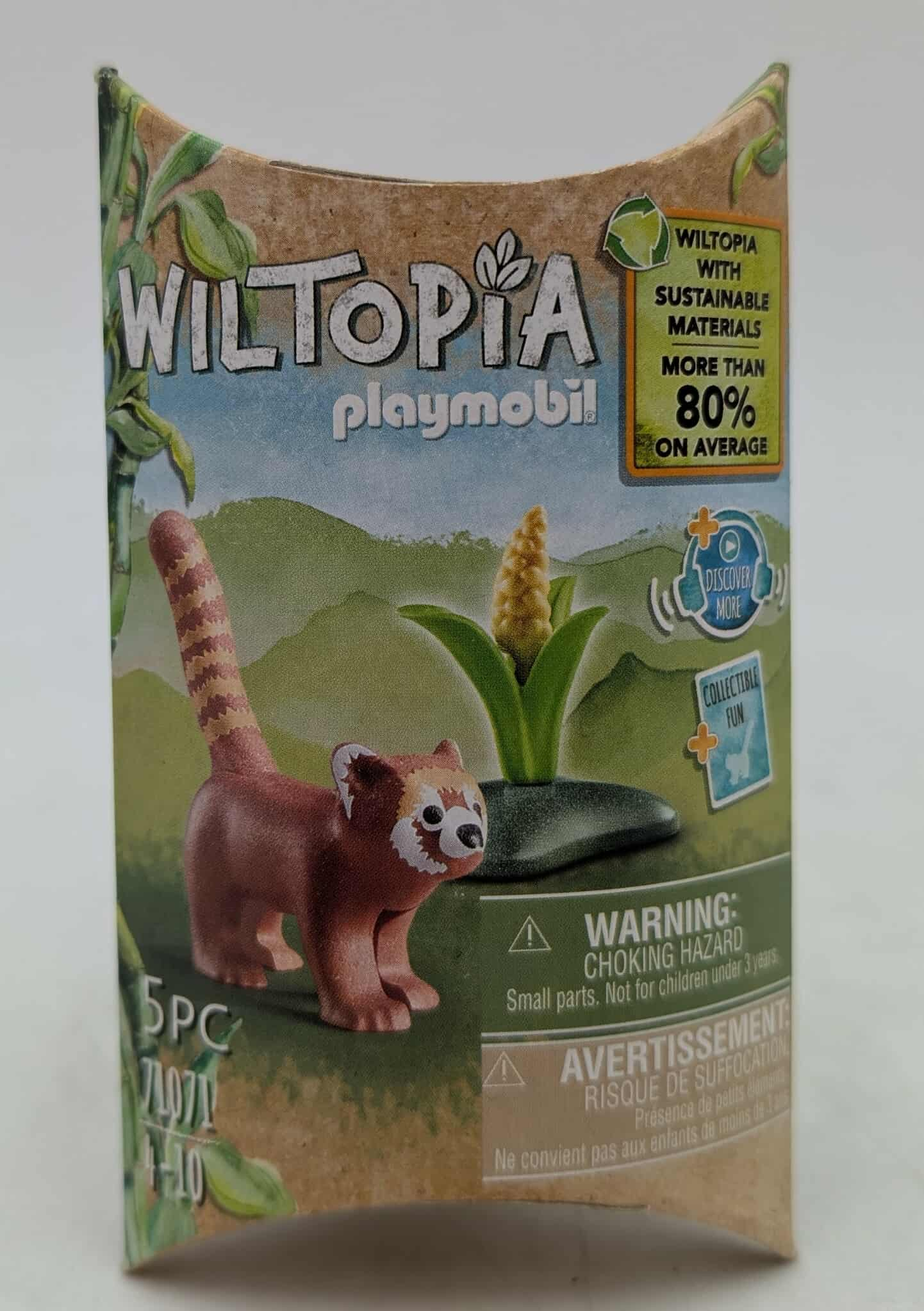 A Playmobil box features a red panda toy. The box displays 'Wiltopia' branding and sustainability claims. The background shows a green landscape.