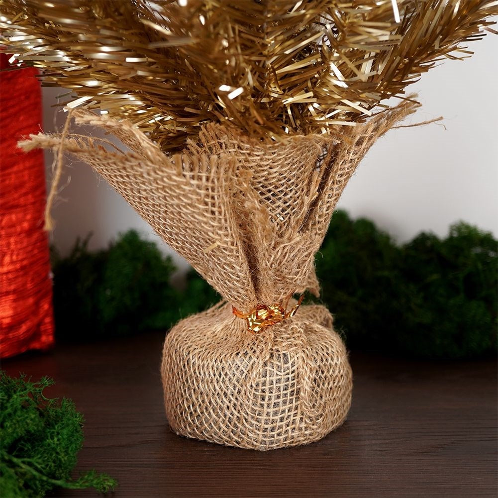 Un pequeño árbol de Navidad dorado envuelto en arpillera. Un objeto rojo a la izquierda. Plantas verdes al fondo. Mesa marrón oscuro.