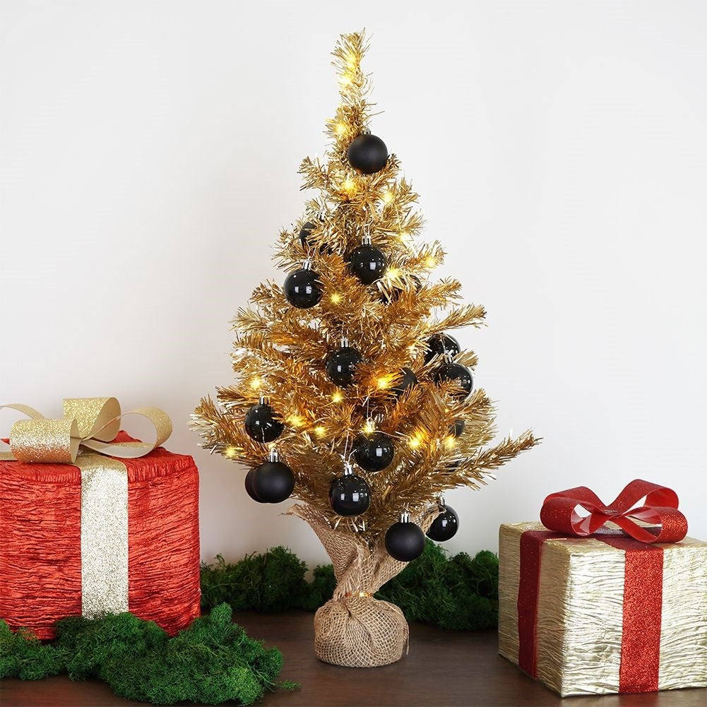 Un árbol de Navidad dorado con adornos negros y luces se encuentra entre dos cajas de regalo. Una caja es roja, la otra dorada.