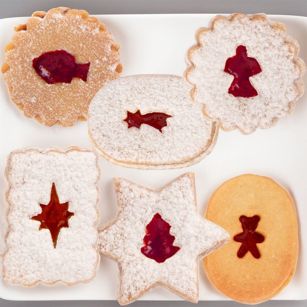 Galletas en plato blanco. Formas diversas: pez, estrella, rectángulo, óvalo, estrella, ángel. Rellenas de mermelada roja, espolvoreadas con azúcar glas.
