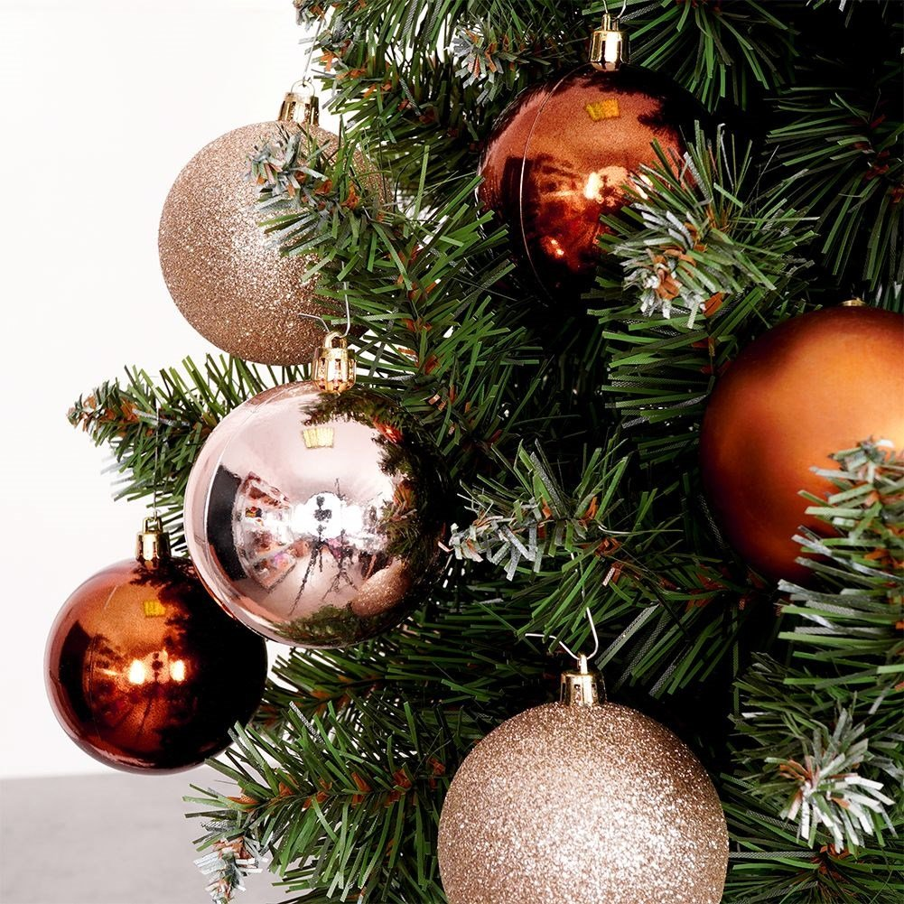 Primer plano de un árbol de Navidad con adornos brillantes. Bolas doradas, bronce y oro rosa cuelgan entre agujas verdes. Fondo blanco.