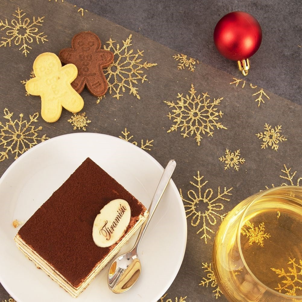 Una rebanada de tiramisú en un plato blanco con una cuchara, galletas, un adorno rojo y un vaso de líquido sobre una mesa decorada.