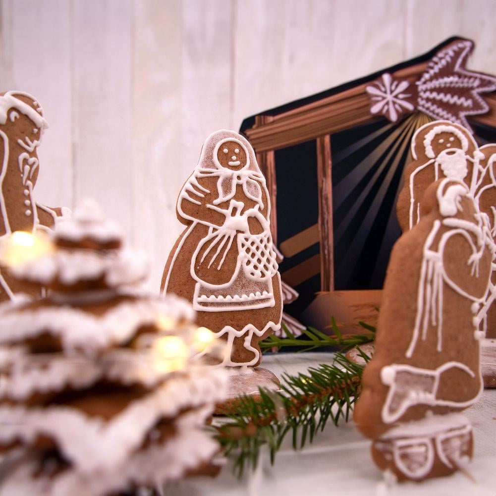 Figuras de pan de jengibre representan una escena de la natividad, incluyendo una mujer, un árbol y un establo. Glaseado blanco detalla las galletas marrones.