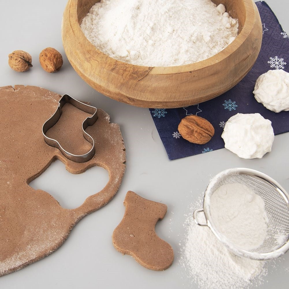 Un cuenco de madera con harina, masa con cortador de galletas en forma de calcetín, nueces, malvaviscos y un tamiz con harina.