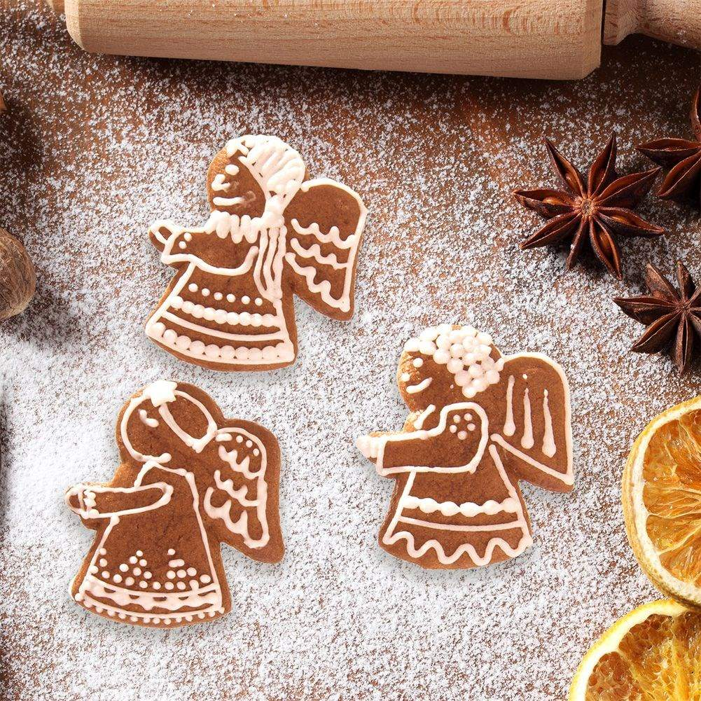 Tres galletas de jengibre con forma de ángel sobre una superficie de madera espolvoreada con azúcar glas, rodeadas de anís estrellado y rodajas de naranja secas.