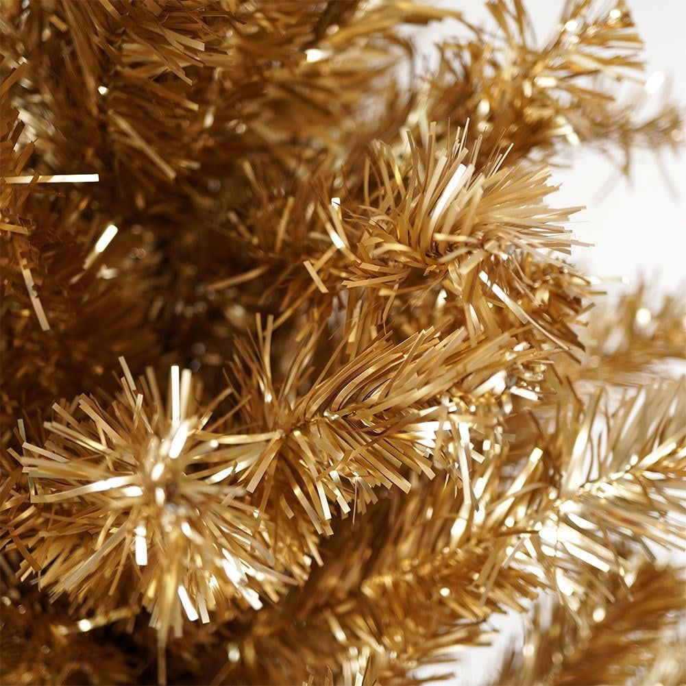 Primer plano de un árbol de Navidad dorado artificial. Las agujas son finas, brillantes y en varios tonos dorados. Fondo blanco.