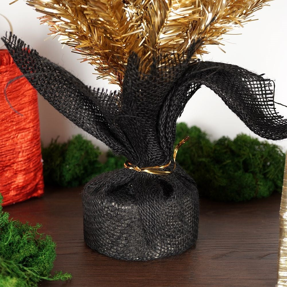 Un pequeño árbol de Navidad artificial dorado está envuelto en arpillera negra y atado con un anillo dorado. Musgo verde en el fondo.
