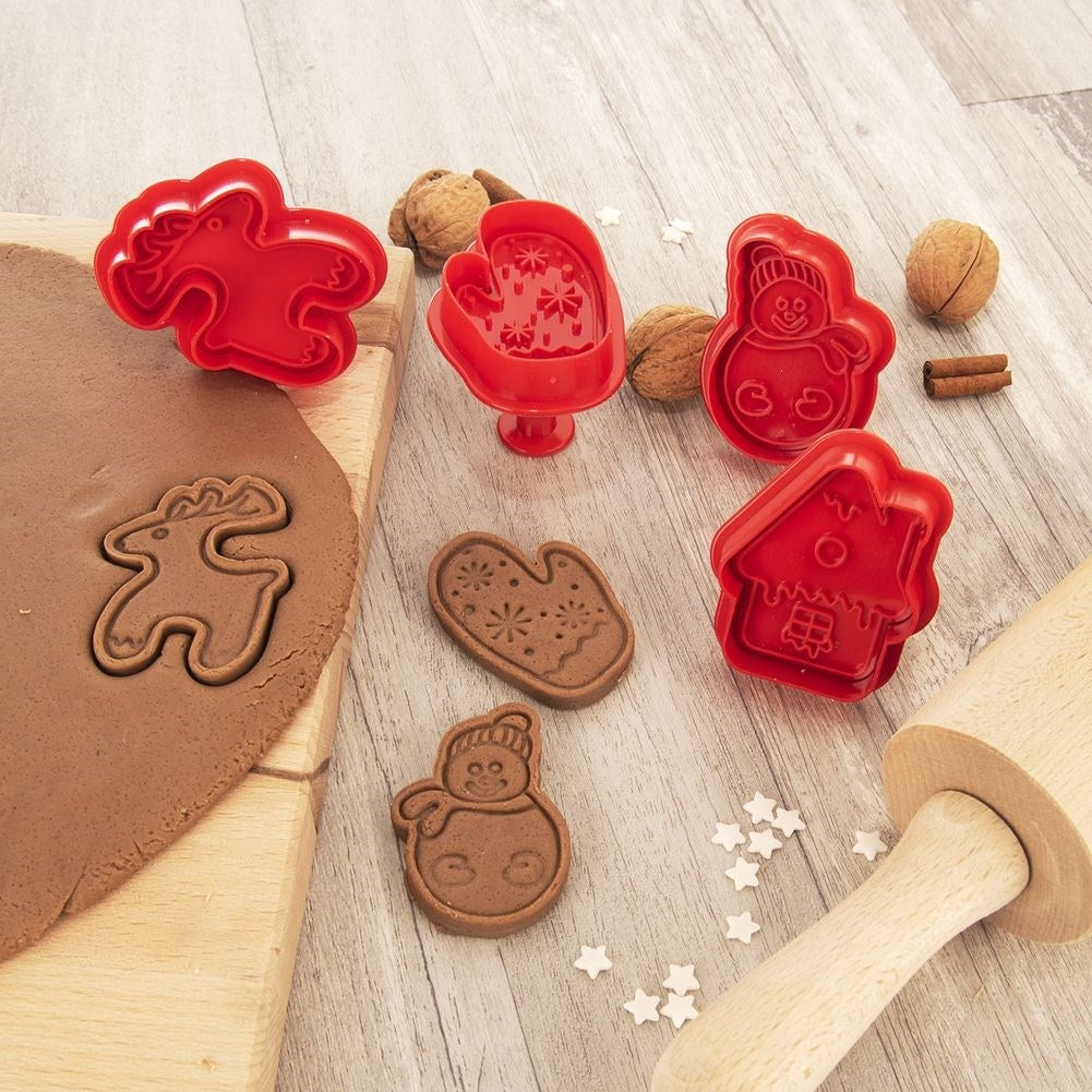 Cortadores de galletas rojos sobre una mesa de madera. Hay masa, galletas, rodillo, nueces y palitos de canela.