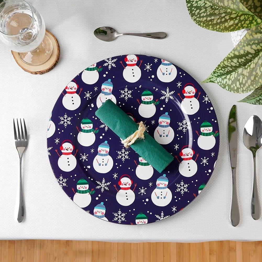 Una mesa con un plato decorado con muñecos de nieve, servilleta verde, cubiertos, vaso y planta. El plato es azul.