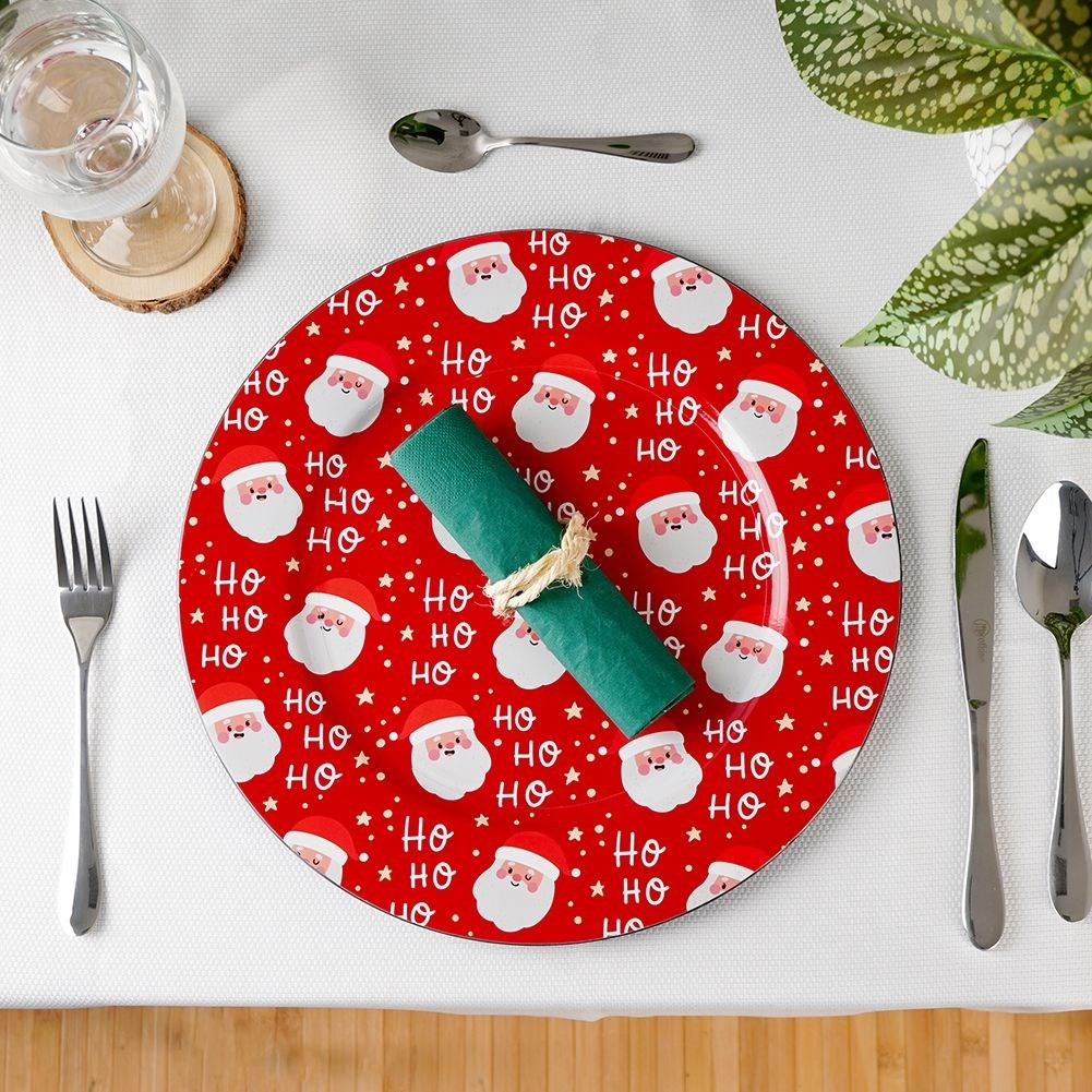 Un arreglo de mesa navideño con un plato rojo con caras de Papá Noel, servilleta verde, cubiertos y un vaso.