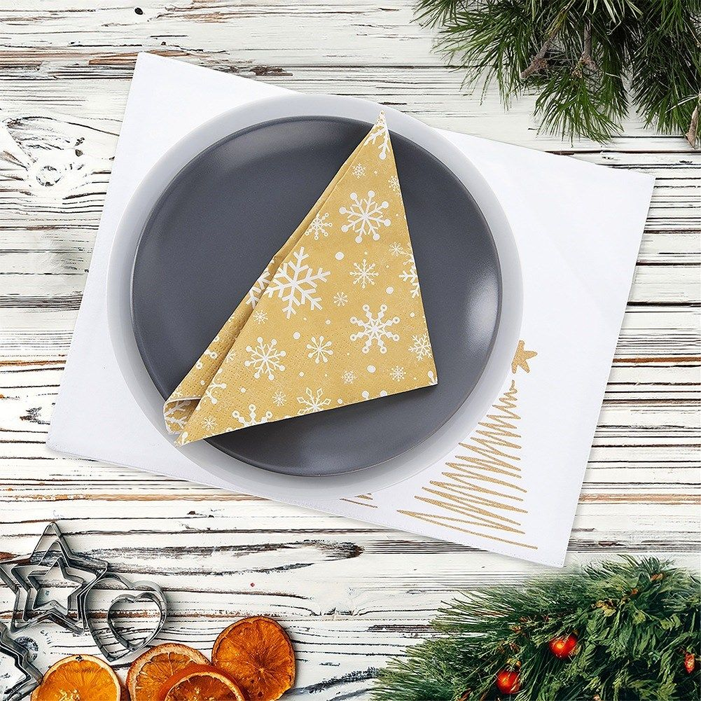 Un montaje de mesa navideño. Servilleta dorada con copos de nieve en plato gris. Mantel blanco con árbol dorado. Naranjas secas en la mesa.