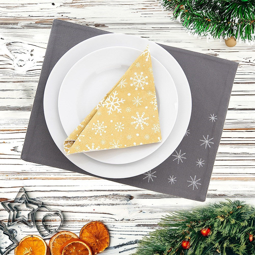 Una mesa navideña con platos, una servilleta de copos de nieve, un mantel individual gris y decoraciones. Se ven naranjas secas y ramas de pino.
