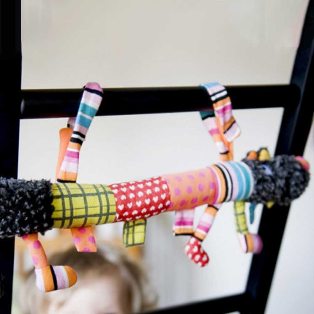 Un juguete de peluche colorido con secciones estampadas y patas colgantes cuelga de un marco negro. La cara de un niño es visible en el fondo.