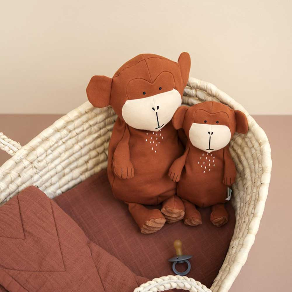 Dos peluches de mono marrón sentados en una cesta de mimbre. Una manta marrón y un chupete están dentro de la cesta. El fondo es beige.