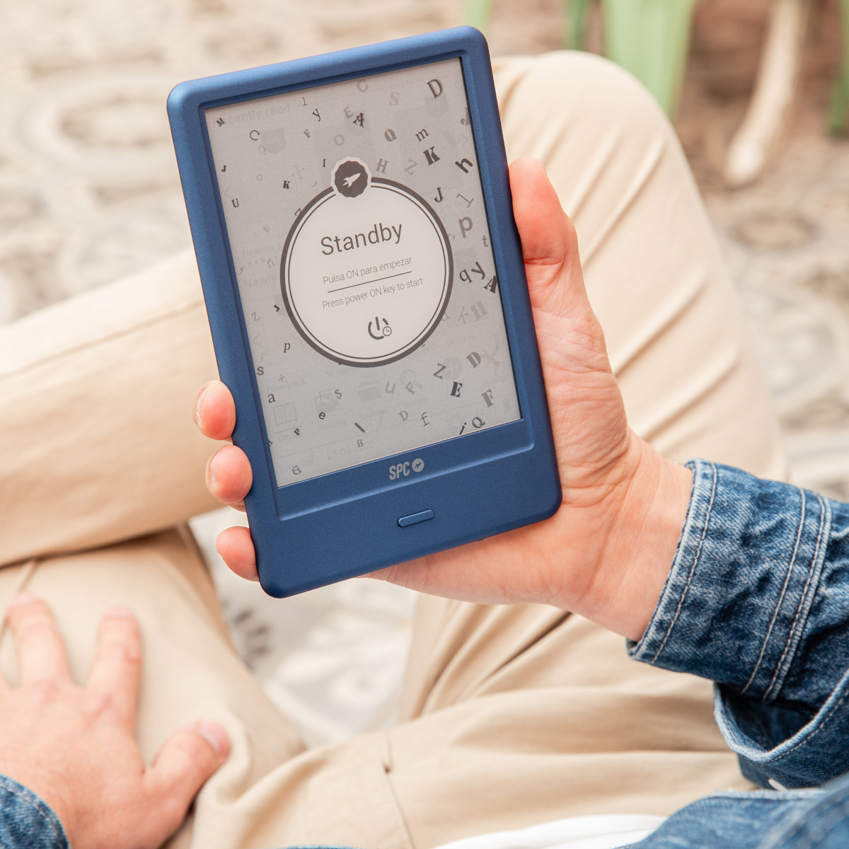 Osoba trzyma niebieski czytnik e-booków z ekranem gotowości. Osoba ma na sobie dżinsową kurtkę i beżowe spodnie.