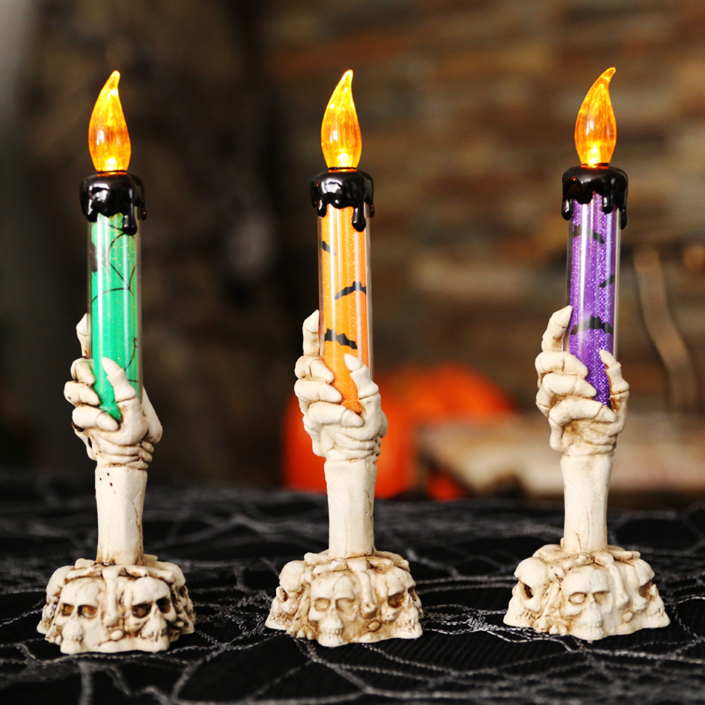 Drei Halloween-Kerzen, die von Skeletthänden gehalten werden. Kerzen sind grün, orange und lila mit schwarzem Wachs. Schädel verzieren die Sockel.