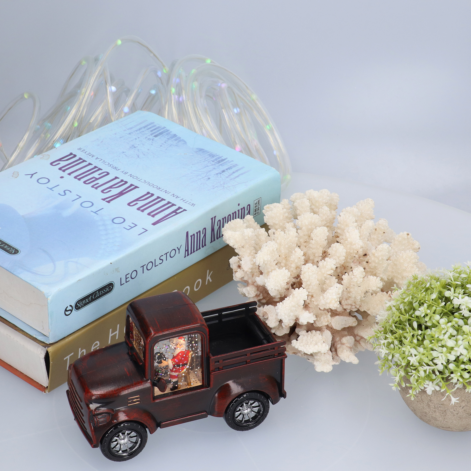 Ein Stillleben zeigt Bücher, einen Spielzeug-LKW mit Santa, Korallen und eine kleine Pflanze. Im Hintergrund sind Lichterketten zu sehen.